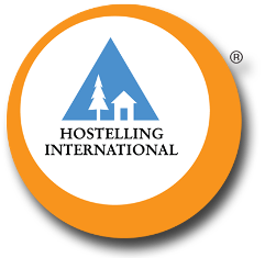 Logotyp organizacji HOSTELS & INSPIRATION w formie pomarańczowego koła wewnątrz któego na białym tle znajduje sie piktogram budynku i drzewa.  